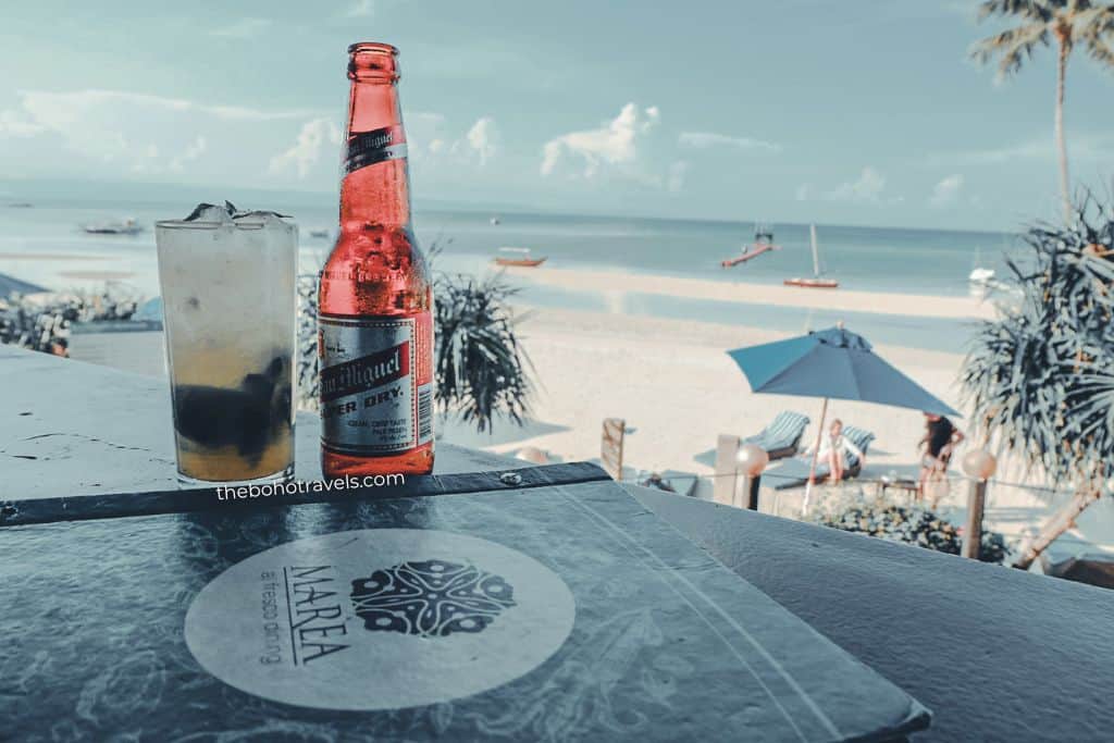 An iced drink and a bottle of beer sit on a table overlooking a white sand beach with boats anchored offshore. Enjoying refreshments with an ocean view is one of the relaxing things to do in Bohol.