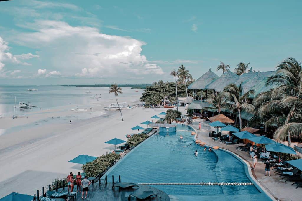 An infinity pool overlooks a white sand beach lined with palm trees, umbrellas, and boats anchored in clear blue water. Relaxing at a beachfront resort is one of the popular things to do in Bohol.