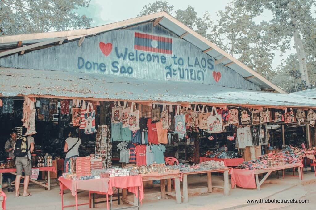 Souvenir stalls at Don Sao Island in Laos with a Welcome to Laos sign, a brief stop on a Chiang Rai day trip from Chiang Mai.