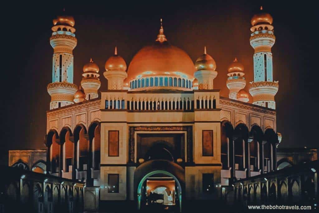 A grand mosque with multiple illuminated minarets and golden domes glows against the dark night sky. Its ornate architecture showcases the distinctive Islamic design found in Brunei.