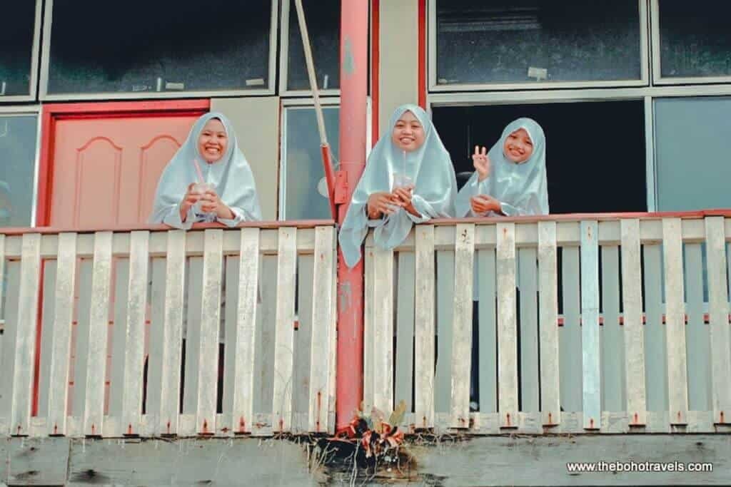 Three young women wearing light colored hijabs smile and wave from the balcony of a waterside building. The photo captures a friendly moment of everyday life in Brunei.
