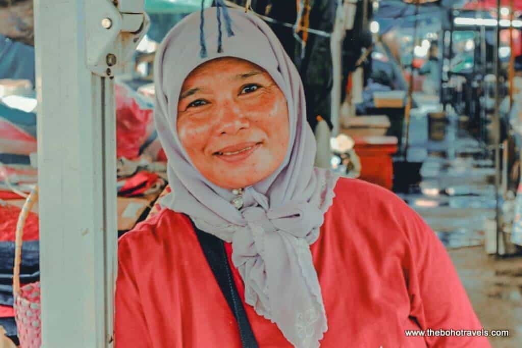 A smiling woman wearing a light gray hijab and bright red top stands at a market stall in Brunei. Her welcoming expression offers a glimpse of the local people and everyday culture travelers can experience when visiting Brunei.