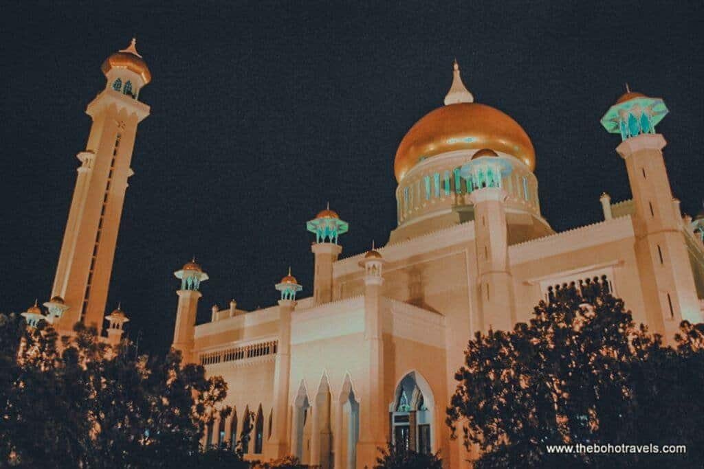 The illuminated Sultan Omar Ali Saifuddien Mosque rises above trees at night, with its large golden dome and tall minarets glowing against the dark sky. The dramatic view highlights one of Brunei's most recognizable landmarks.