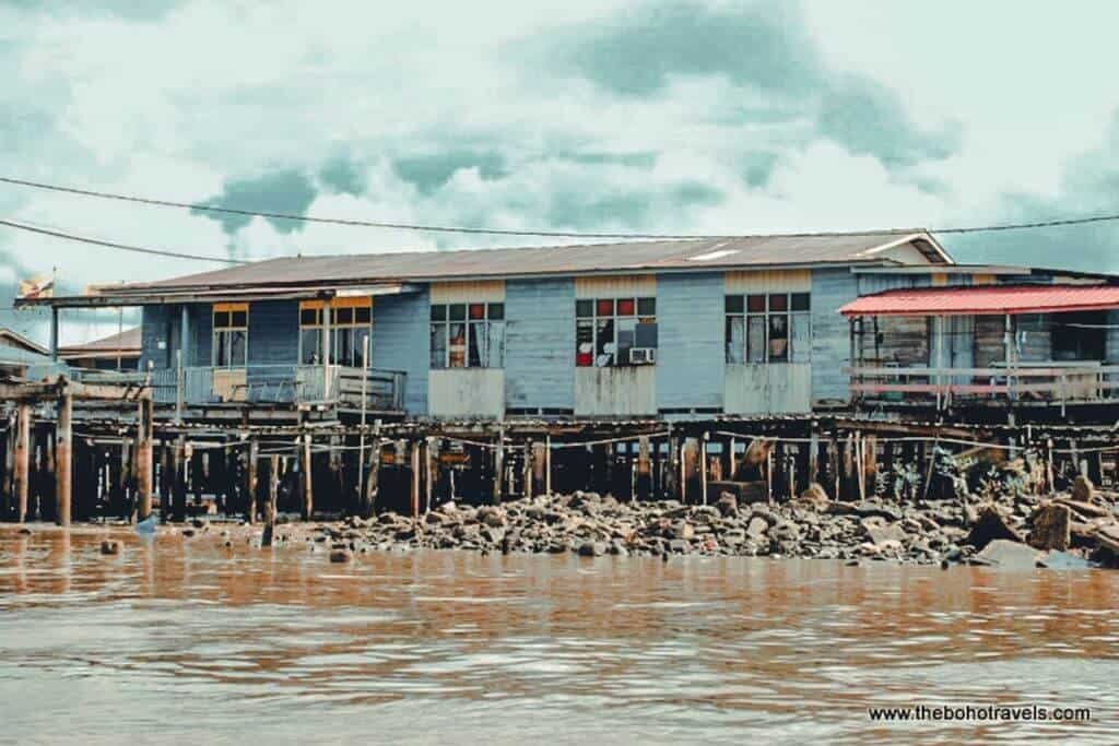 A weathered wooden house stands on stilts above the brown waters of the Brunei River, with rocky shoreline beneath it. The traditional waterfront homes provide a glimpse into life around Kampong Ayer.