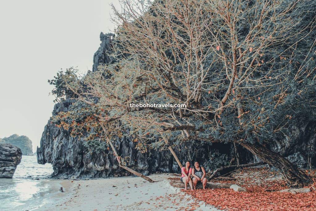 Two girls in one of the islands of El Nido, resting under the tree with sand between their feet, answering YES to the question - is El Nido worth visiting