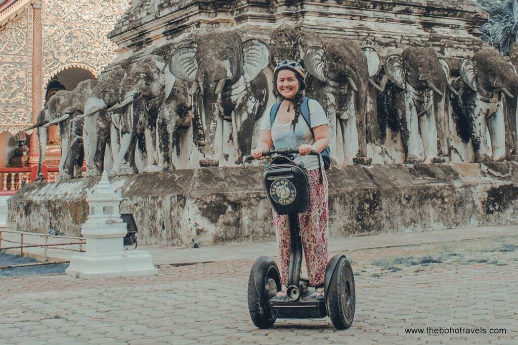 A woman rides a Segway in front of a historic row of elephant statues at a temple complex in Chiang Mai. The scene captures a fun and unique way to explore cultural attractions and things to do in Chiang Mai.