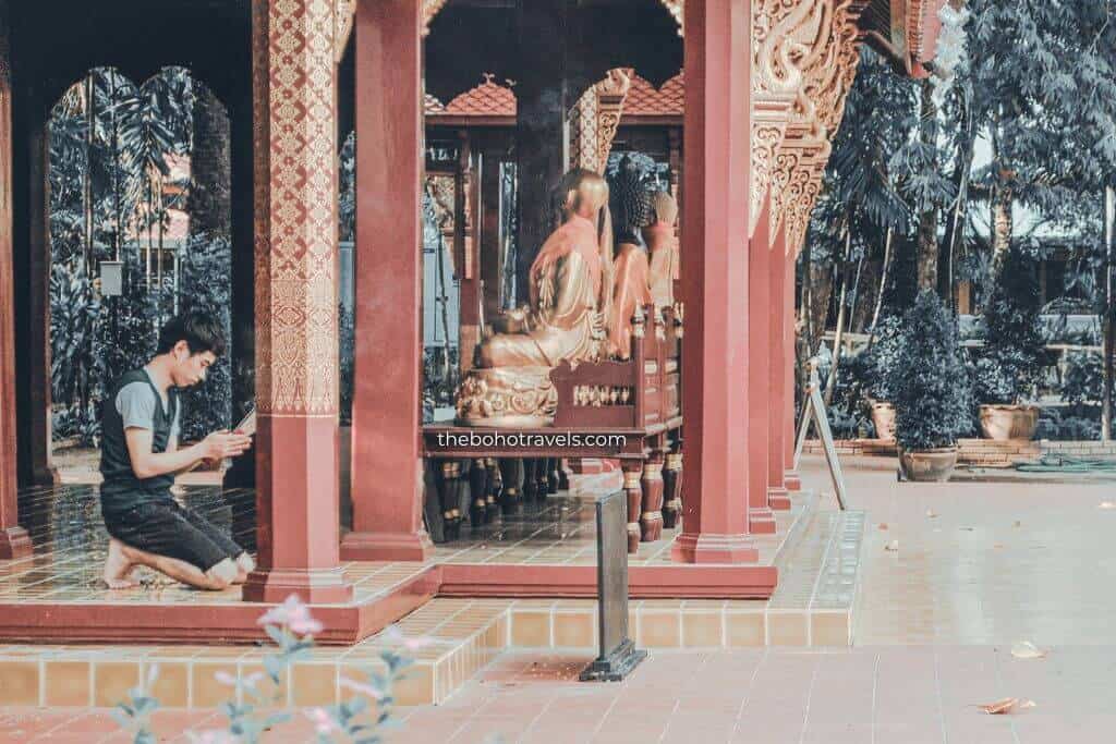 A man kneels in prayer beside golden Buddha statues inside an ornate temple in Chiang Mai. The peaceful temple setting highlights the cultural and spiritual side of things to do in Chiang Mai.