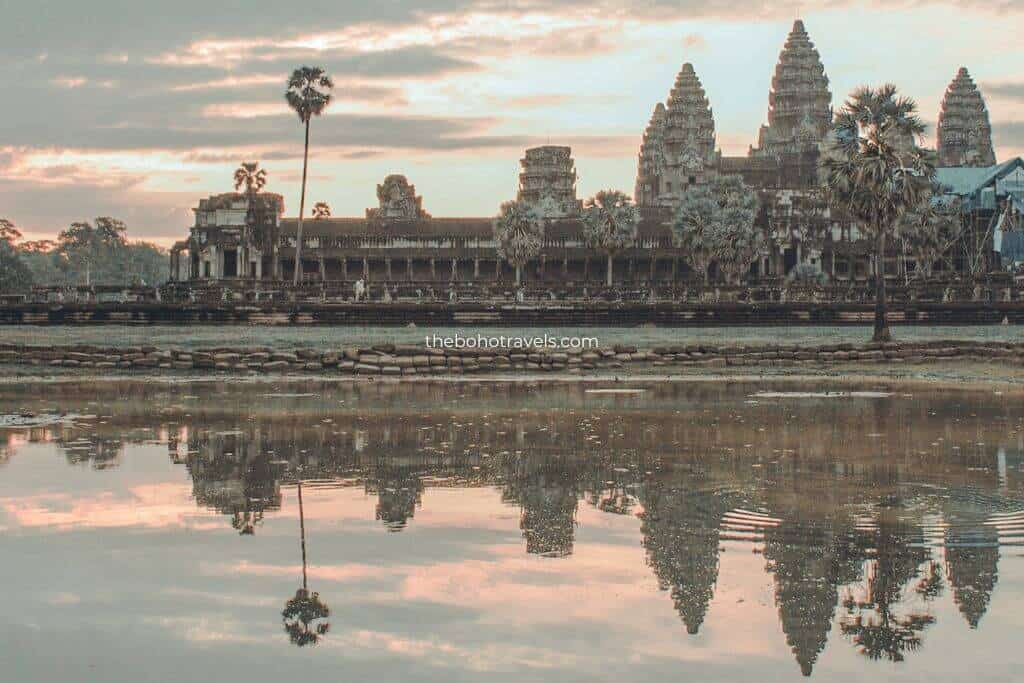 An ancient temple complex rises behind a calm reflecting pond at sunrise, with palm trees silhouetted against the colorful morning sky. The peaceful scene showcases one of the region's most iconic archaeological sites featured in a Southeast Asia travel guide.