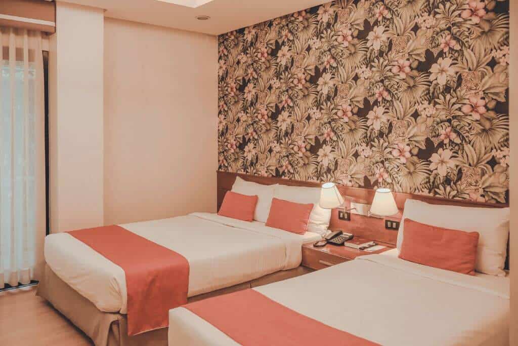 Two neatly made beds with coral accent pillows and runners sit against a bold floral feature wall inside a guest room at Giraffe Boutique Hotel in Baguio. Warm lighting, wooden furnishings, and a large window create a cozy and inviting space for travelers staying in the city.