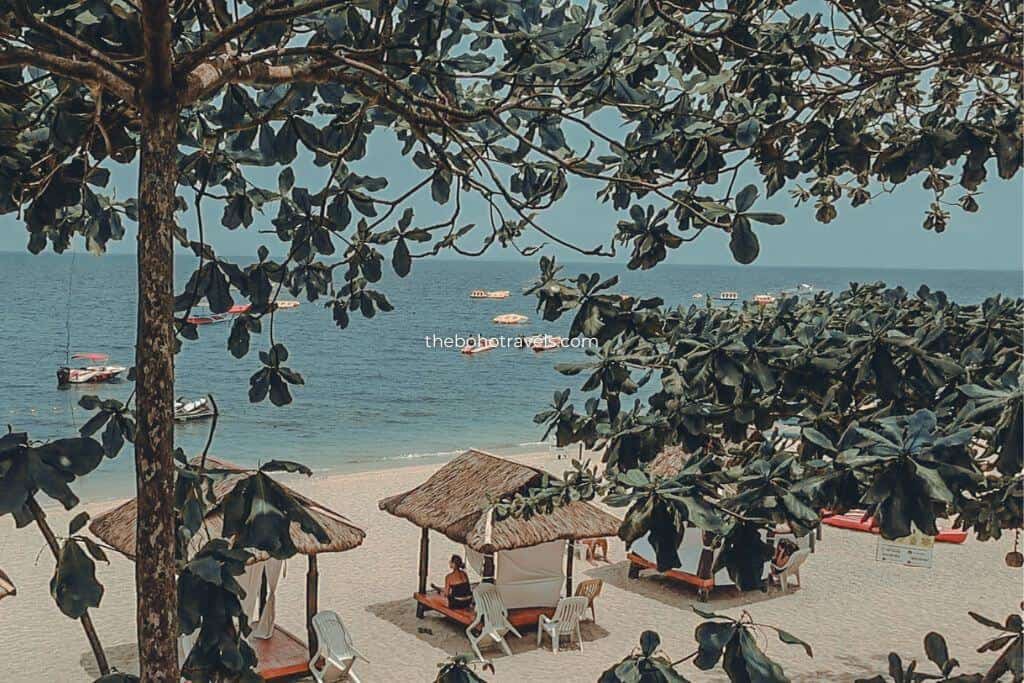 Elevated view of a sandy beach lined with thatched cabanas and shaded by large coastal trees. Calm blue water with small boats anchored offshore creates a relaxing seaside atmosphere at The Henry Resort Taramindu Laiya, featured in this The Henry Resort Taramindu Laiya Review.