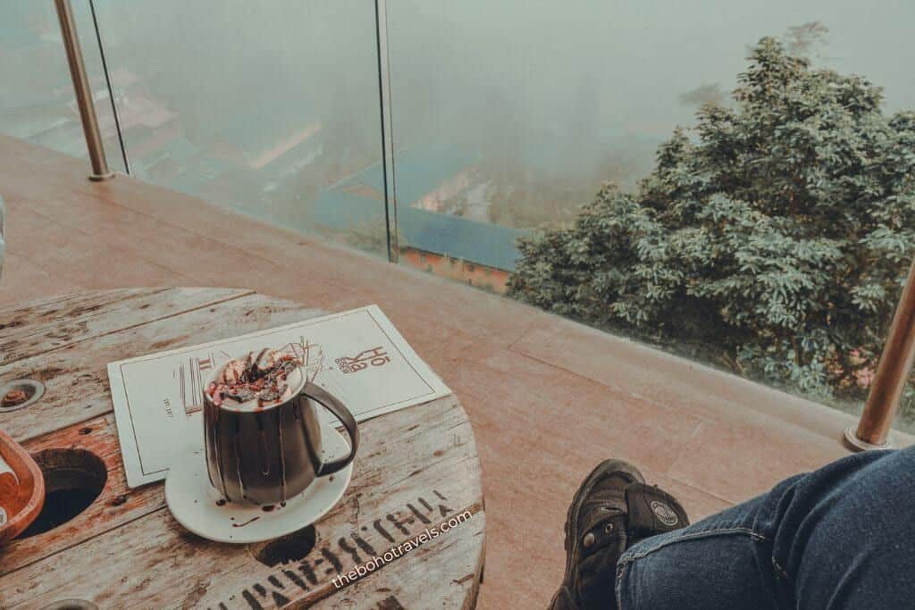 Outdoor seating area at Hoka Brew, located at the 2nd Floor of The Podium Boutique Hotel, with a mug of hot chocolate topped with whipped cream and chocolate drizzle resting on a rustic wooden table. A glass railing overlooks misty hillsides and lush trees, creating a peaceful mountain view.