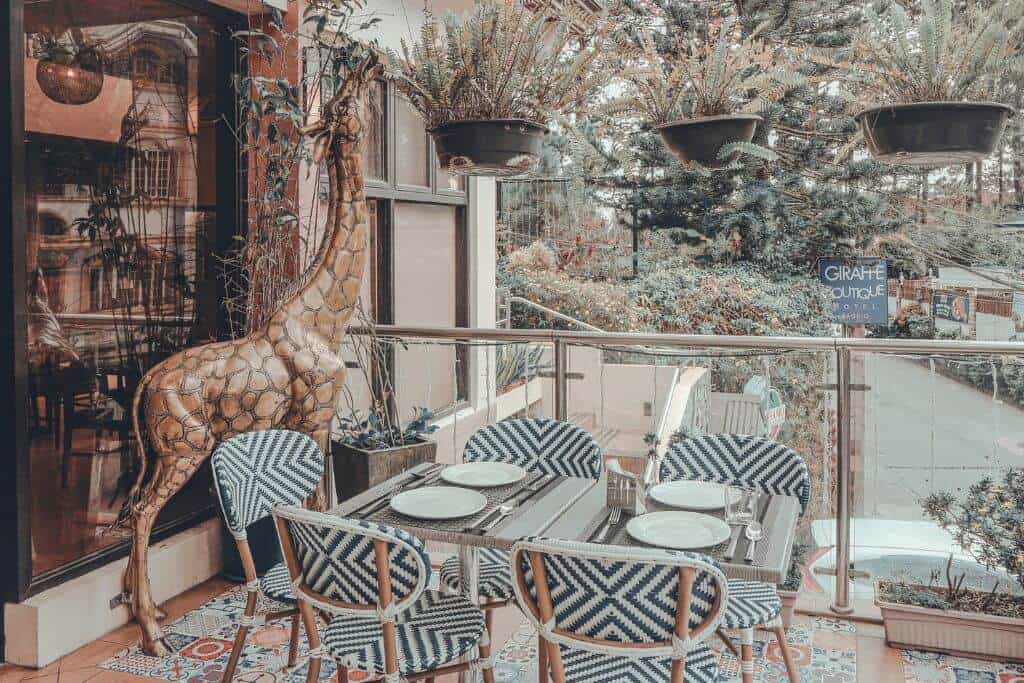 Outdoor dining area at Giraffe Boutique Hotel in Baguio featuring patterned chairs, a table set with plates, hanging plants, and a decorative giraffe sculpture beside the entrance. The terrace overlooks a tree lined street, creating a relaxed garden atmosphere for guests dining or gathering outdoors.