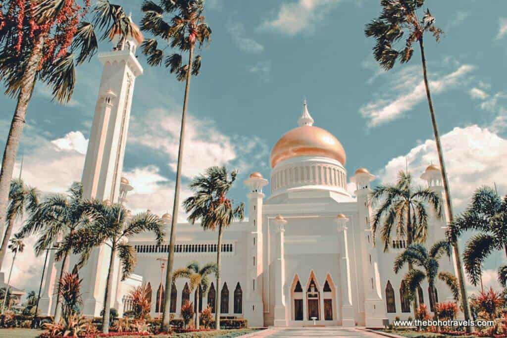 A white mosque with a large golden dome and tall minaret stands among palm trees beneath a bright blue sky with scattered clouds. The striking architecture reflects the cultural and religious diversity highlighted in a Southeast Asia travel guide.