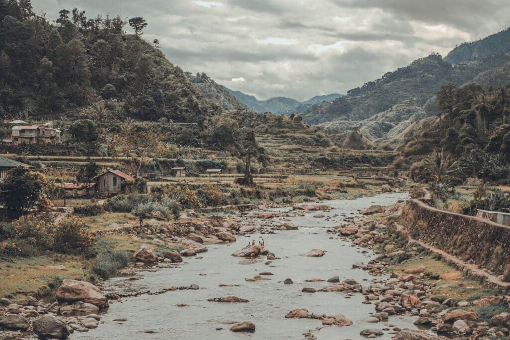 A rocky river winds through a mountain valley lined with terraced fields, scattered homes, and dense forested hills. Low clouds hang over the distant mountains, highlighting the rural landscape and layered terrain.