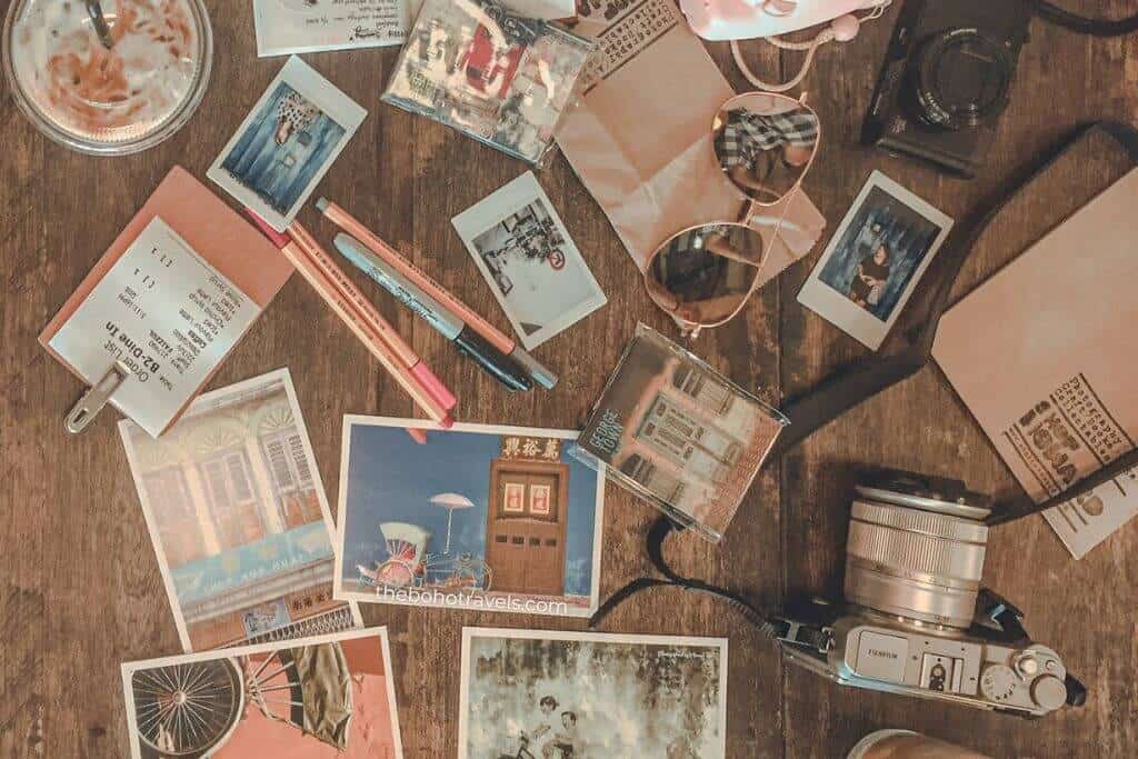 A flat lay of travel essentials including postcards, instant photos, a camera, sunglasses, colorful pens, a passport, and an iced coffee arranged on a wooden table. The collection captures the excitement of planning and documenting adventures throughout a Southeast Asia travel guide.
