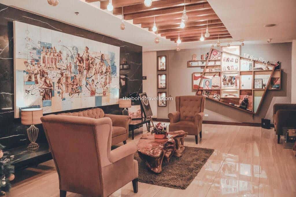 Lounge area inside The Podium Boutique Hotel featuring plush armchairs, a natural wood coffee table, warm pendant lighting, and decorative wall displays. A large mural and cozy seating arrangement create an inviting space for guests to relax.