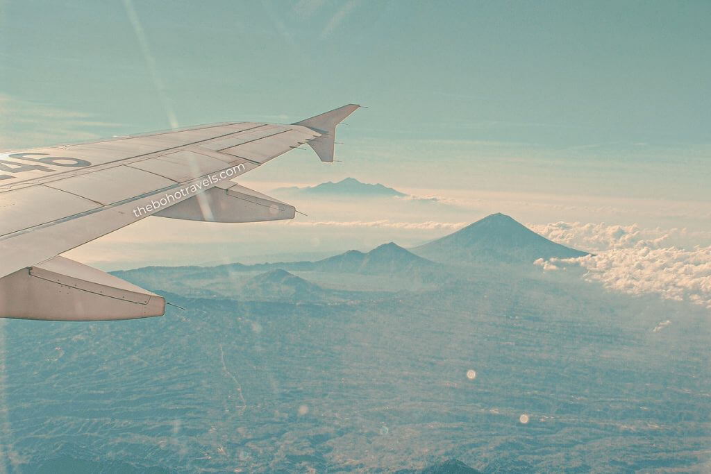 An airplane wing extends above a layer of clouds with volcanic peaks rising from the landscape below. The aerial view captures the excitement of arriving in the region and the dramatic scenery travelers can experience throughout a Southeast Asia travel guide.