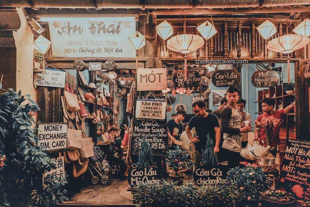 A bustling street-side restaurant is filled with hanging lanterns, handwritten menu boards, and diners enjoying local cuisine in a lively market setting. The vibrant food scene highlights the authentic flavors and cultural experiences that make dining a memorable part of a Southeast Asia travel guide.