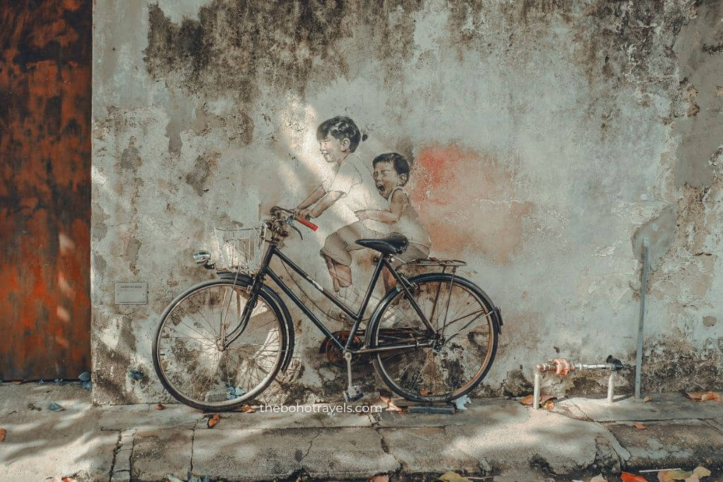 A vintage bicycle is parked in front of a weathered wall featuring a mural of two children riding together, creating the illusion that the bicycle is part of the artwork. This playful piece of street art reflects the creative urban culture and unique photo spots often featured in a Southeast Asia travel guide.