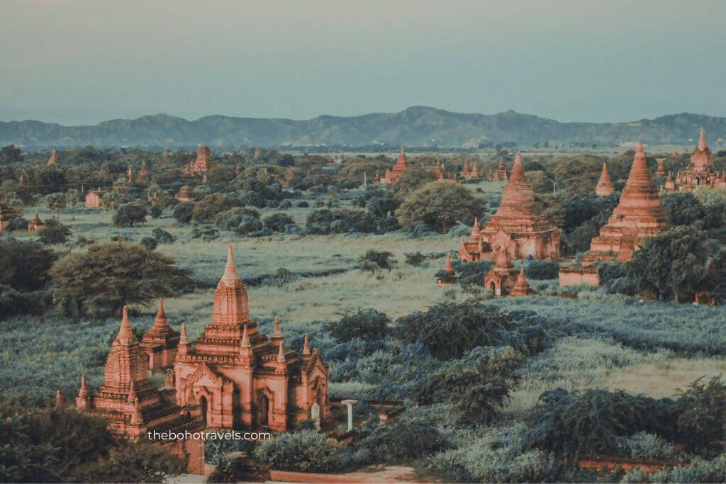 An expansive landscape of ancient brick pagodas and temples stretches across a green plain with mountains rising in the distance under a clear sky. The historic archaeological site showcases the rich cultural heritage and unforgettable landmarks featured in a Southeast Asia travel guide.