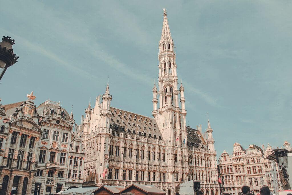 The ornate Gothic architecture of Brussels Town Hall rises above the Grand Place under a blue sky, surrounded by historic guild houses. This landmark showcases the impressive architecture and cultural heritage featured in a Europe travel guide.