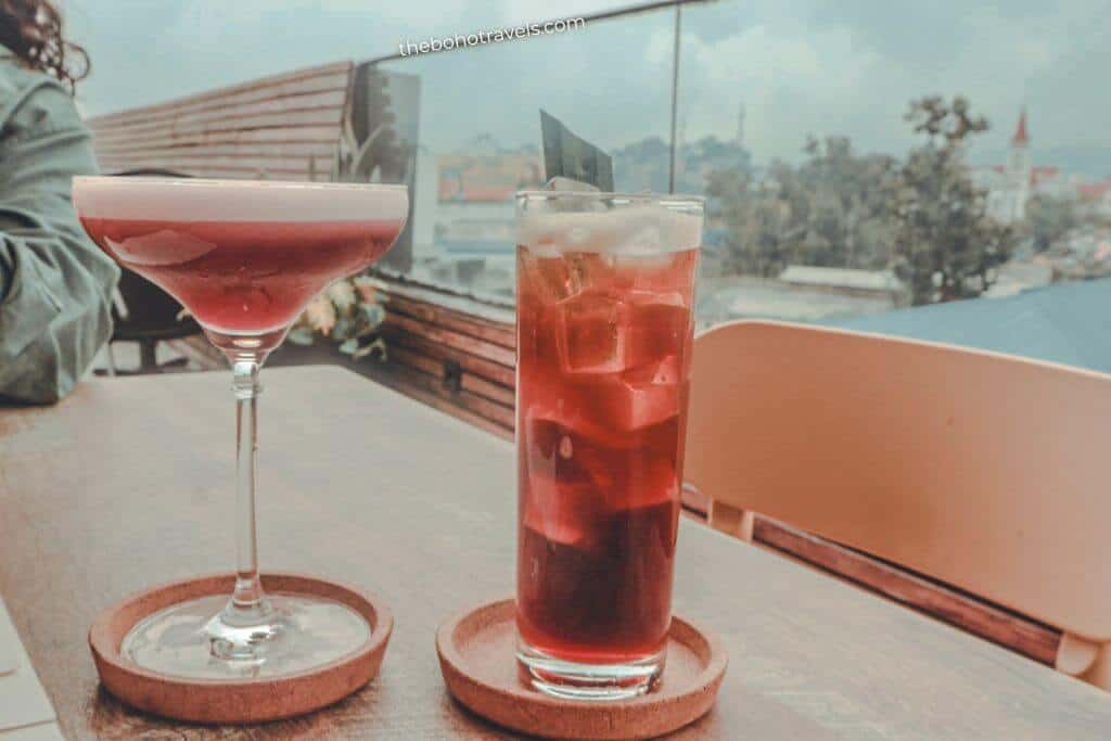 A pink cocktail in a coupe glass and a tall iced mixed drink rest on wooden coasters beside a window overlooking the city. The drinks and scenic view make this a relaxing stop for where to drink in Baguio.