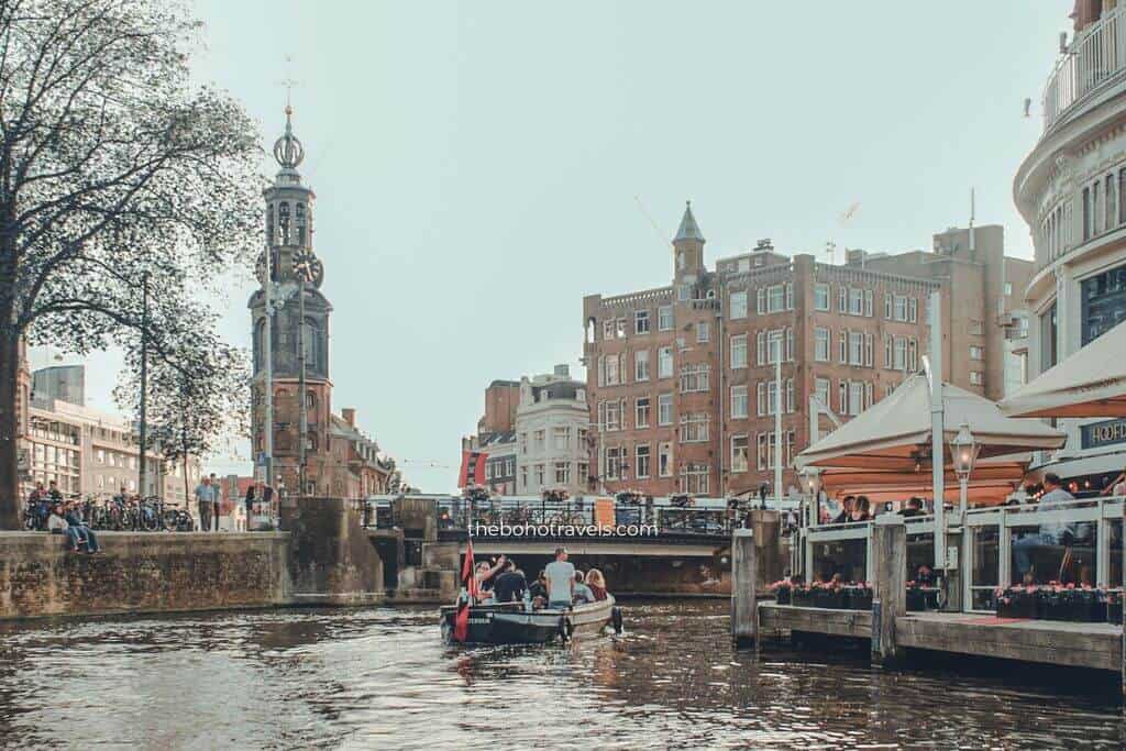 A small boat carrying passengers cruises along an Amsterdam canal toward the Munttoren clock tower, with waterfront cafés and historic buildings lining the water. This scene captures the relaxed atmosphere that makes the city a memorable destination in a Europe travel guide.