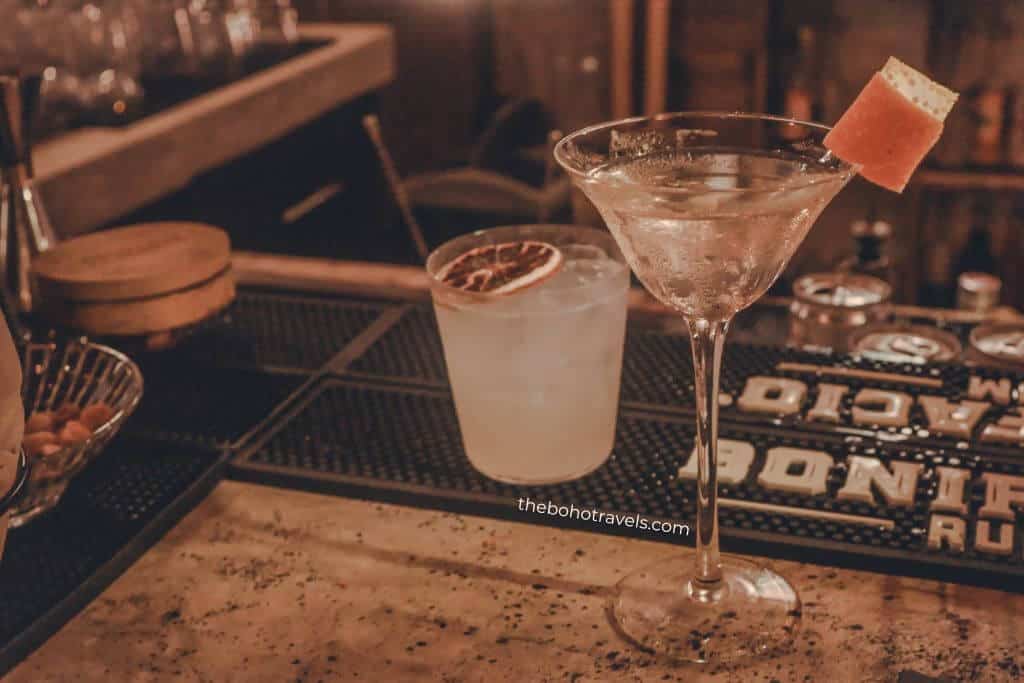 Two handcrafted cocktails sit on a bar counter, including a martini garnished with a grapefruit peel and a short citrus drink topped with a dried orange slice. These signature drinks showcase where to drink in Baguio for creative cocktails.