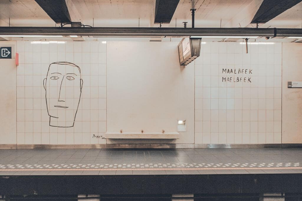 The Maalbeek metro station platform features a minimalist line drawing of a face on a tiled wall beside the station name "MAALBEEK MAELBEEK." The quiet platform highlights a recognizable stop in Brussels that travelers may encounter while following a Europe travel guide.