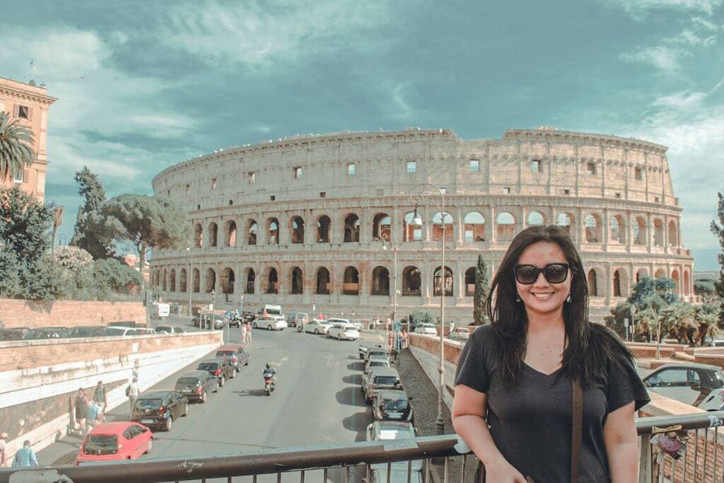 A woman in blue wearing shades in front of the Colosseum in Rome, towering above a busy street with cars, pedestrians, and trees beneath a partly cloudy sky. This famous ancient amphitheater is one of the most recognizable landmarks featured in a Europe travel guide.