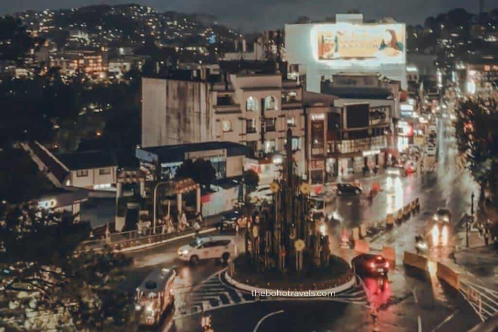 Nighttime view of a busy city roundabout with glowing streetlights, moving traffic, and illuminated buildings after the rain. The vibrant cityscape captures the evening atmosphere before a night out in Baguio.