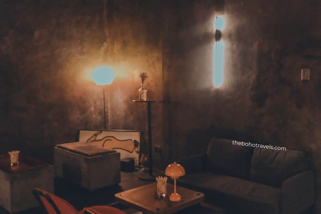 Cozy lounge area with low sofas, warm ambient lighting, textured walls, and modern decor creates a relaxed setting for drinks and conversation. The moody interior highlights a hidden speakeasy in Baguio.