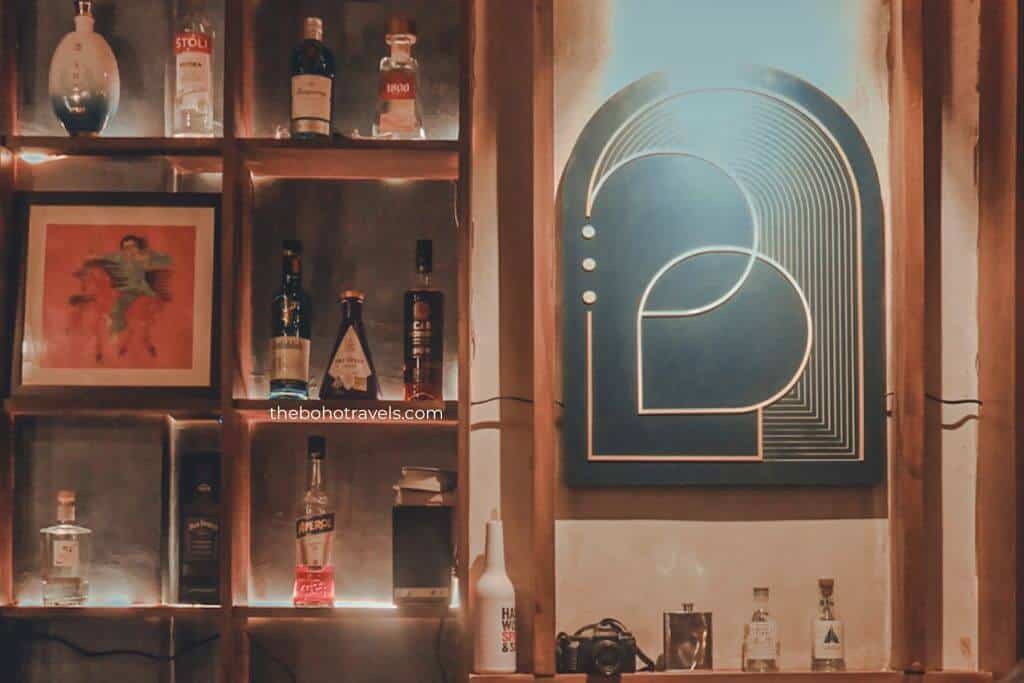 Backlit shelves display bottles of liquor, framed artwork, and bar decor beside a glowing geometric wall light in a dimly lit cocktail bar. The stylish interior reflects the intimate atmosphere of a speakeasy in Baguio.