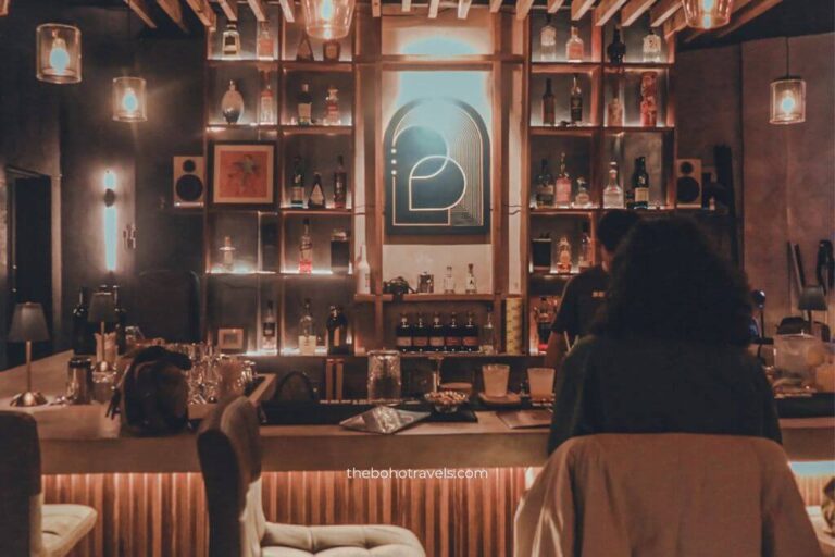 Warmly lit cocktail bar with shelves of liquor bottles, hanging pendant lights, and guests seated at the bar creating an intimate lounge atmosphere. This cozy interior captures the ambiance of speakeasies in Baguio, making it a great choice for travelers looking for where to drink in Baguio.