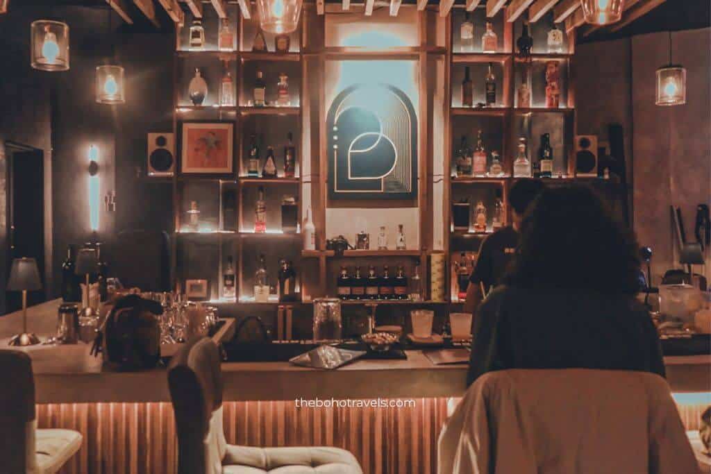 Warmly lit cocktail bar with shelves of liquor bottles, hanging pendant lights, and guests seated at the bar creating an intimate lounge atmosphere. This cozy interior captures the ambiance of speakeasies in Baguio, making it a great choice for travelers looking for where to drink in Baguio.