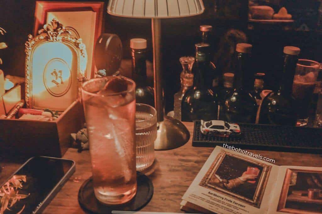 A cocktail served over ice sits on a wooden bar beside vintage decor, liquor bottles, and a softly glowing table lamp. The intimate setting reflects the cozy atmosphere of speakeasies in Baguio, making it an inviting stop for anyone deciding where to drink in Baguio.