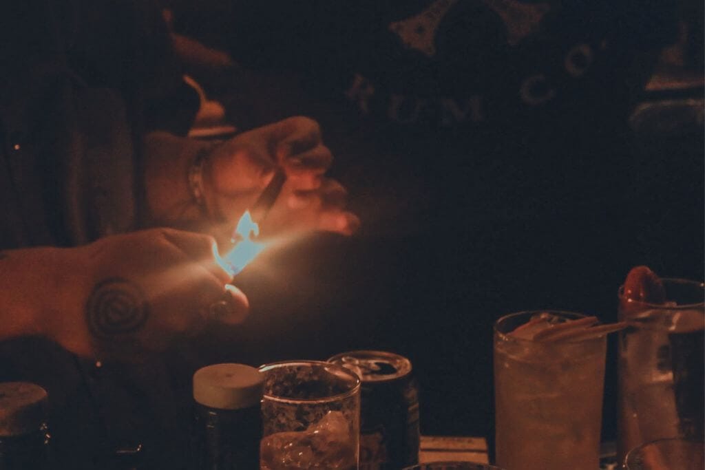 A bartender lights a garnish over handcrafted cocktails, adding a dramatic finishing touch in a low-lit bar. The cocktail preparation highlights the creative drinks and immersive experience that make speakeasies in Baguio a memorable answer to where to drink in Baguio.
