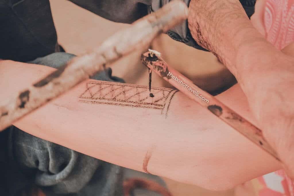 Close up of a person using a bamboo stick to hand apply a traditional tattoo design onto another person's forearm. The process captures a unique cultural experience often documented in travel storytelling.