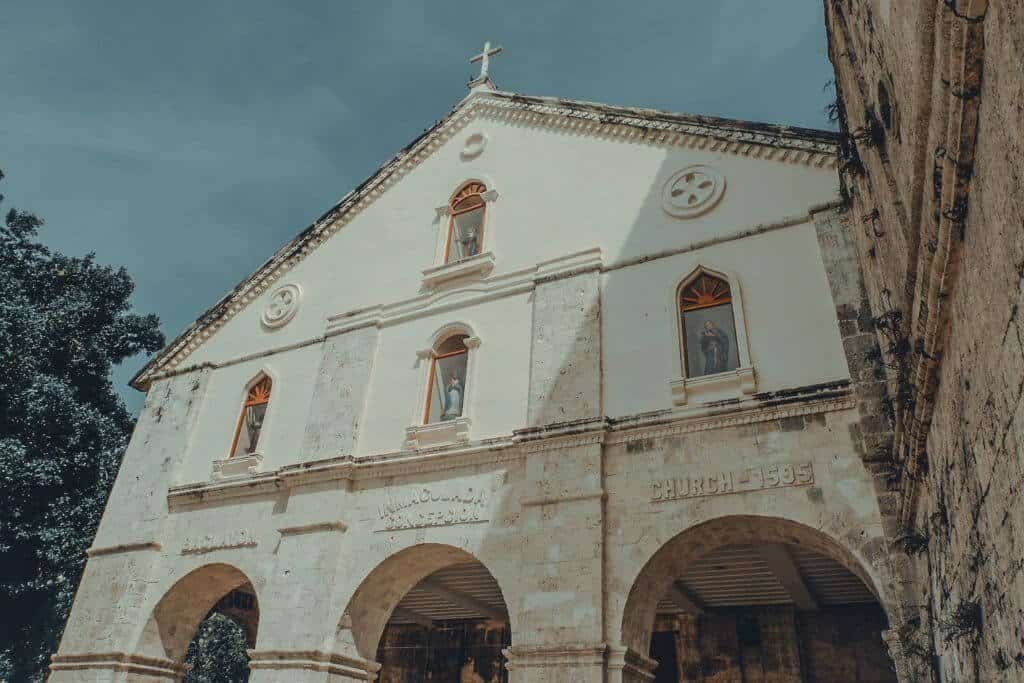 Historic stone church in Bohol with a weathered white facade, arched entrances, religious statues in tall windows, decorative details, and a cross above the roof. The preserved church is one of the cultural and heritage focused Bohol tourist spots.