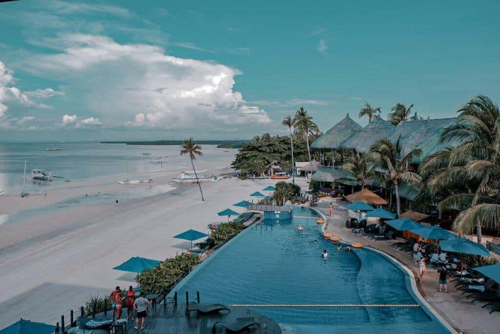 Aerial view of Bellevue Resort in Bohol, a tropical beachfront resort with a large swimming pool overlooking a wide sandy beach and calm blue water, with palm trees, thatched structures, boats, and colorful umbrellas along the shore.
