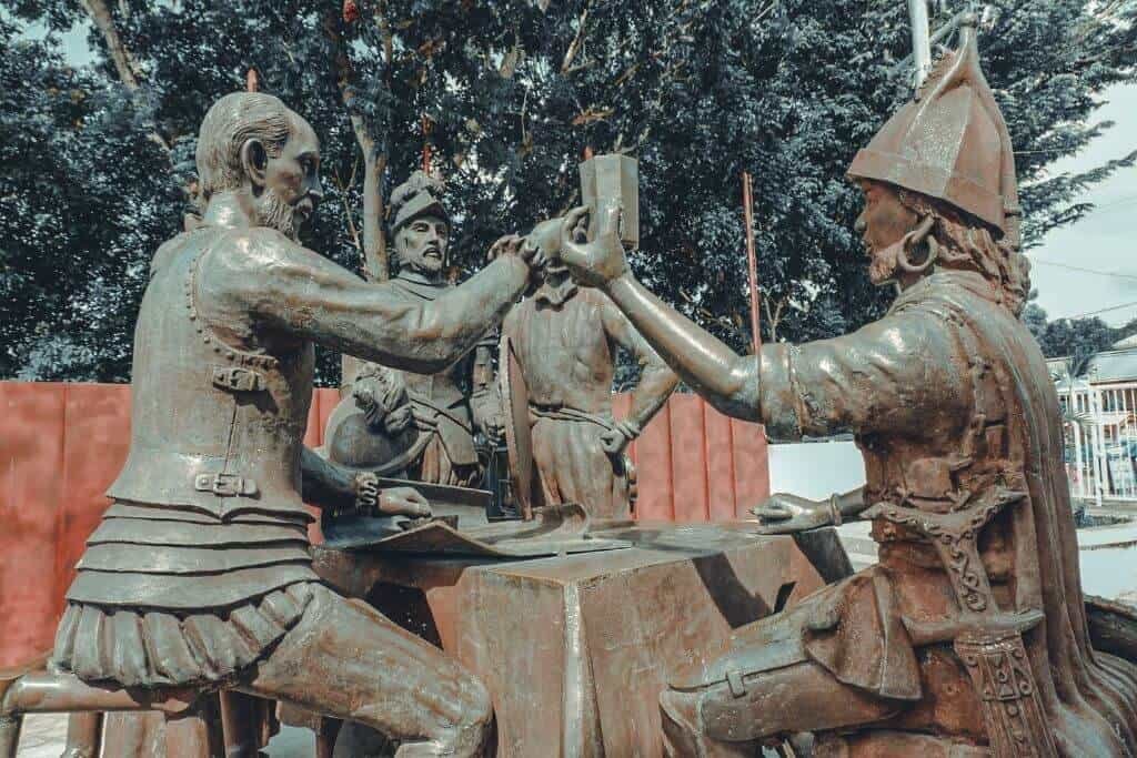 Bronze historical monument showing several figures gathered around a table and raising a cup or vessel, surrounded by trees and buildings. The sculpture represents an important moment in Bohol history.