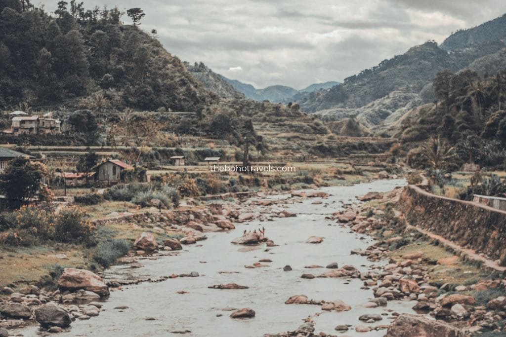 Rocky river winding through a lush mountain valley with scattered homes, terraced fields, and dense forest beneath a cloudy sky. The peaceful rural landscape showcases the kind of scenic destination that inspires travel photography and storytelling.