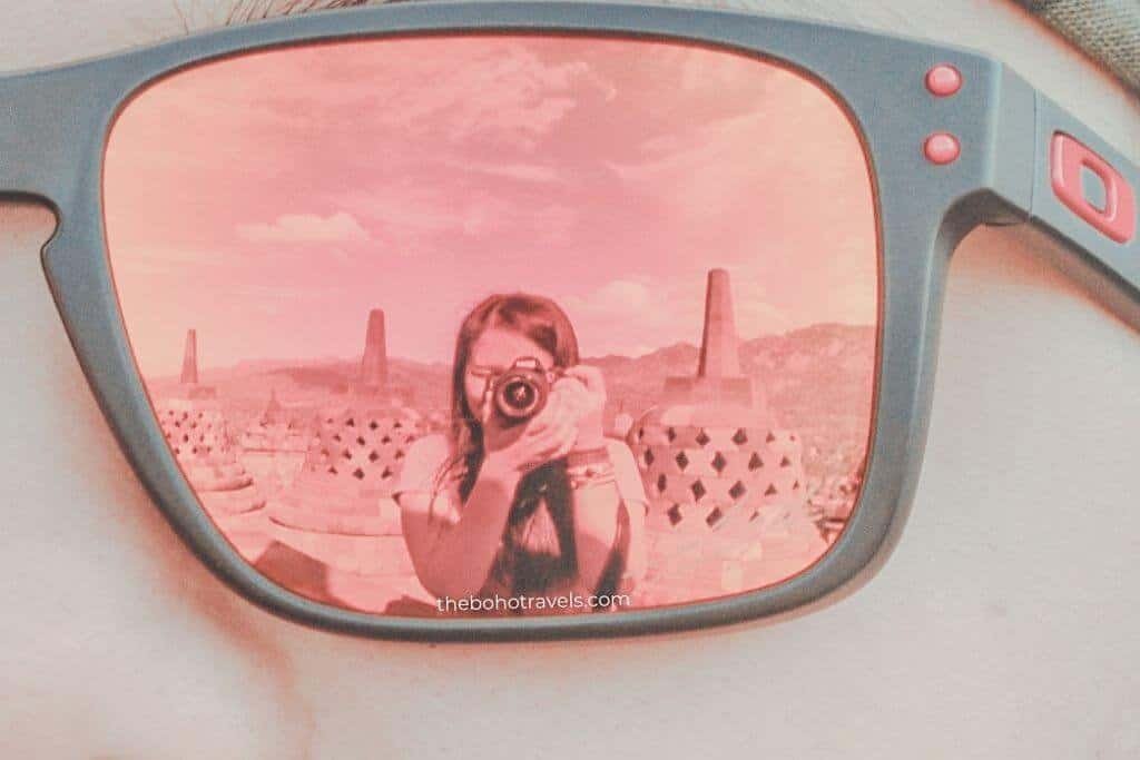 Reflection of a person holding a camera captured in the red tinted lens of a pair of sunglasses, with temple shaped structures visible in the background. The creative self portrait highlights travel photography techniques that can make a travel blog more engaging.