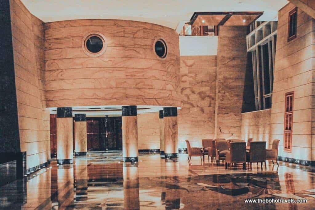 Spacious hotel lobby with polished marble floors, wood toned walls, round windows, and a seating area with upholstered chairs. The warm and formal interior shows the style of accommodation available when deciding where to stay in Brunei.