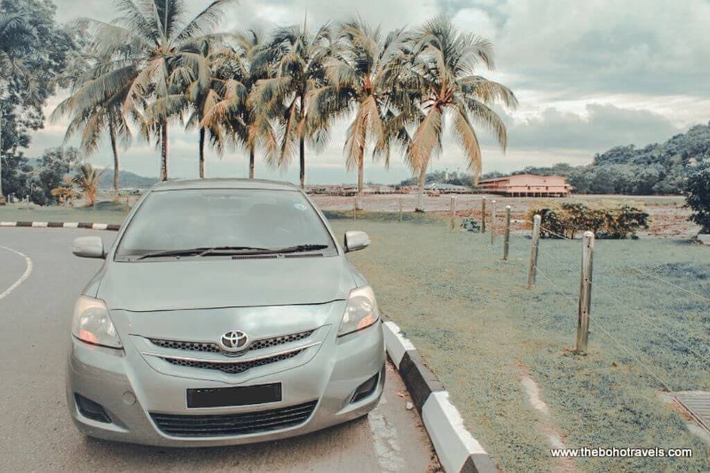 A silver Toyota parked beside a palm lined road with a tropical landscape and low buildings in the background. The setting suggests a hotel or resort in Brunei and provides a glimpse of the surroundings when considering where to stay in Brunei.
