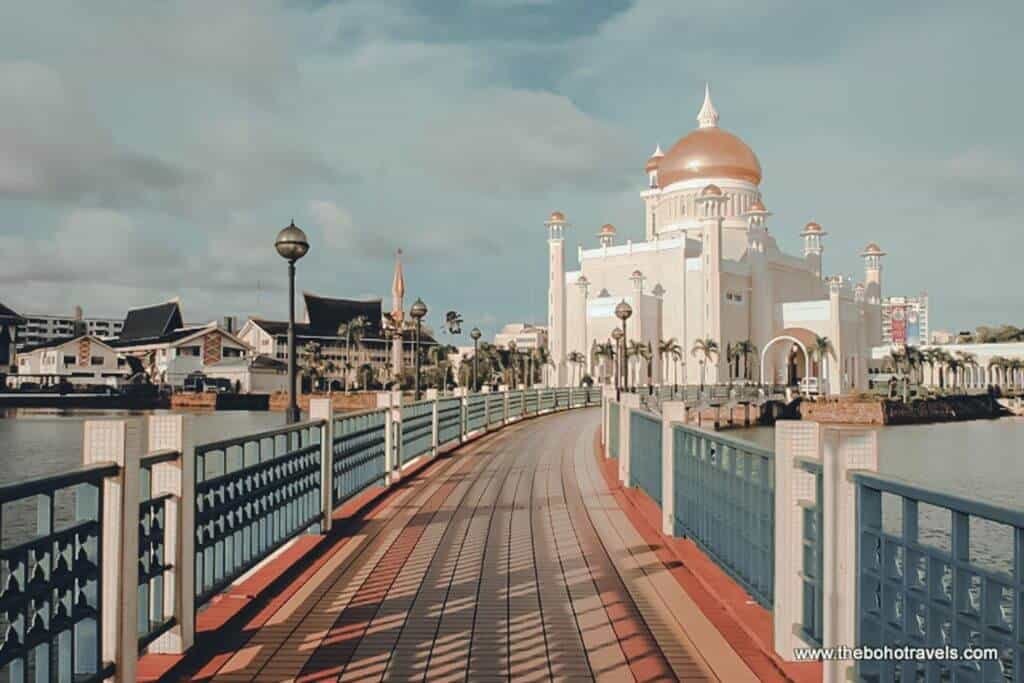 A pedestrian bridge leads toward a large white mosque with a golden dome and ornate minarets beside the water. The landmark and surrounding waterfront provide a view of the Brunei setting for travelers planning where to stay in Brunei.