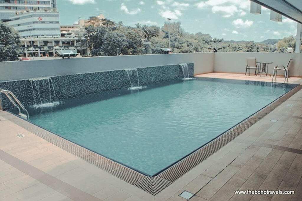 Outdoor hotel swimming pool with a tiled waterfall feature along the far edge and lounge seating overlooking a lush tropical landscape. The pool offers a look at the leisure facilities available at this Brunei accommodation.