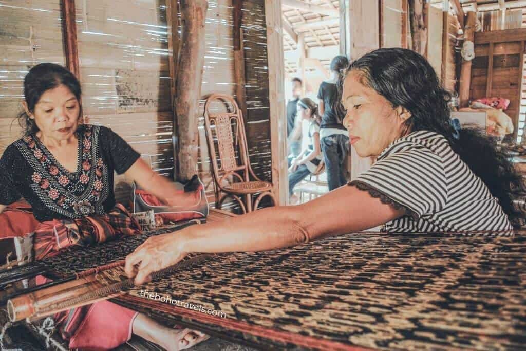 Two women work together on a traditional handloom inside a wooden home, carefully weaving patterned fabric by hand. Learning about local weaving traditions adds cultural depth for anyone researching how to start a travel blog with meaningful travel experiences.