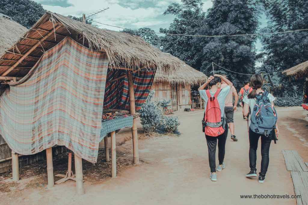 Tourists walking through a Kayan village near the Golden Triangle during a Chiang Rai day trip from Chiang Mai, with bamboo stilt houses and handwoven textiles on display