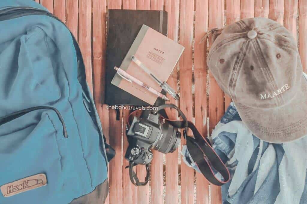 Blue backpack, DSLR camera, notebook labeled "NOTES," two pens, and a gray cap embroidered with "MAARTE" arranged neatly on a bamboo floor. These essential travel and photography items are commonly packed when learning how to start a travel blog.